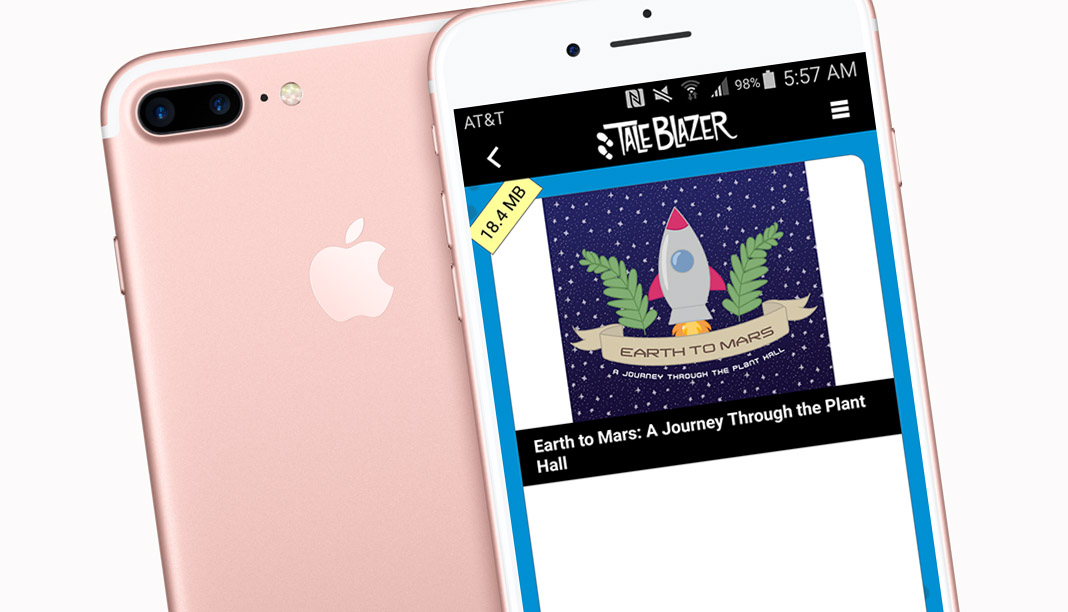 A photo mockup of the game app TaleBlazer. In the photo is an early model pink iPhone, with a second iPhone to its side, with the screen facing up. The screen has a screenshot showing. At the top of the screenshot is a bar with various phone icons, including the battery indicator, the time (5:58), and the Bluetooth icon. Below that is a title reading "TaleBlazer," with icons of two foot prints beside it. Below that is a small yellow diagonal banner that says "18.4 MB." Beside that is a color illustration that shows a blue sky with cartoon stars on it, a red and gray 50's-style cartoon rocket, and two fern leaves on either side of the rocket. Below the rocket is a beige banner that reads "Earth to Mars" in all caps, and below that, the text "A Journey Through the Plant Hall," also in all caps. Below this illustration is a black bar that repeats that title.