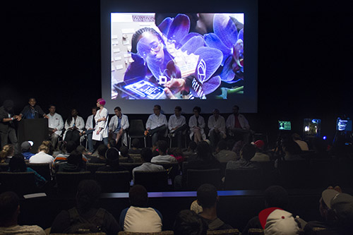Orientation for SEED. Actors, dressed in lab coats, sit in a line on stage. A spotlight shines on a woman with pink hair, also in a lab coat. Some high school students' backs are visible in the audience. Behind the actors a video is playing. A Black girl and boy look at an iPad screen. A purple image appears to be fading in or out of the video.