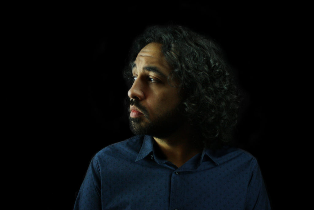 Keith S. Wilson, facing left. Keith has facial hair, brown skin, and medium length curly hair. He is wearing a blue dress shirt.