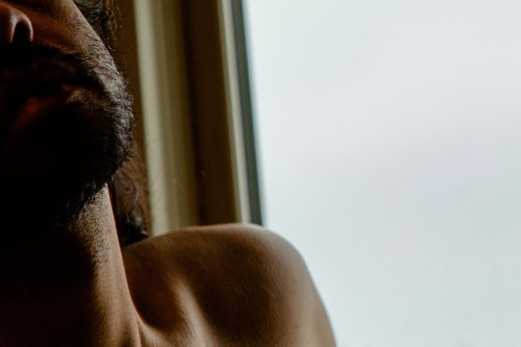 A very close-up photo of a brown skinned man's bearded chin, stubbled neck, and bare shoulder. In the background a white-lit window.