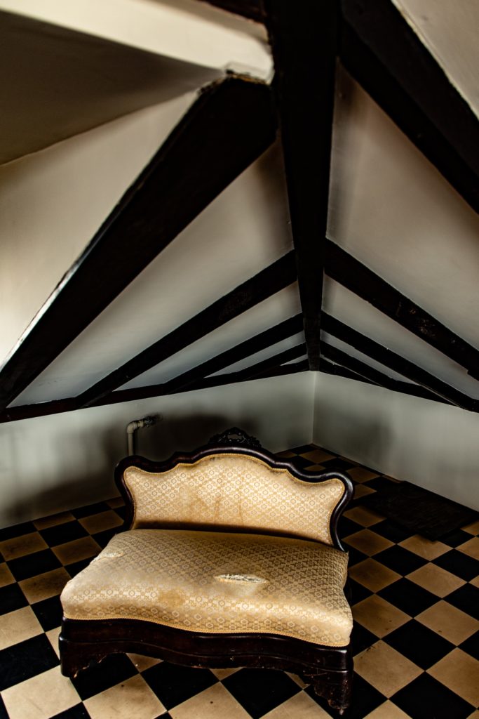 A photo, taken from a high angle, of an old-fashioend torn beige settee sitting in the corner of the room. The floor is dirty black and white checkered linoleum. The white wall appears very short. The wall on the left has an exposed white painted pipe. From the top of the image coming down are the black diagonal beams of the rafters.