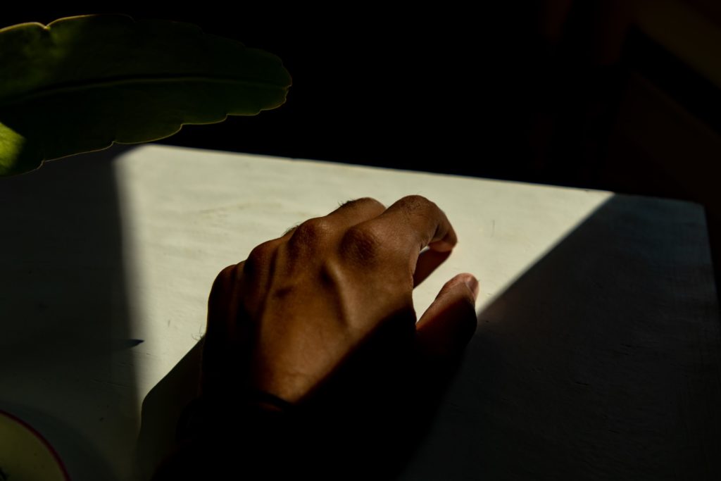 A photograph of a brown hand in triangular light. All else is in shadows too dark to see, besides a green leaf glowing at its edges on the left side of the photo.