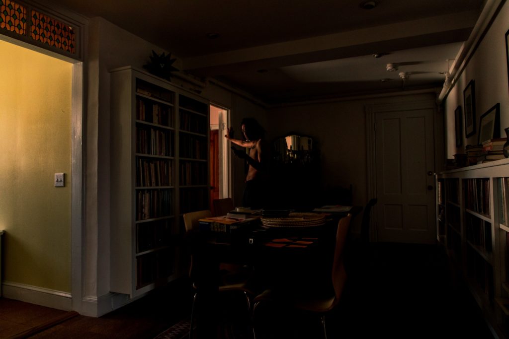 A dark photo of an apartment. The left side of the photo shows the entrance to another room with a yellow painted wall and white light switches. The main room contains a very tall bookcase on the left, and a shorter one on the right, both filled with books. The right-most wall has hanging photos. The center of the room has a long dining table with plates, a puzzle, and other papers on it. At the far back of the room is a mirror, barely lit. Standing in the doorway to another room is a brown-skinned man, shirtless, looking at the camera.