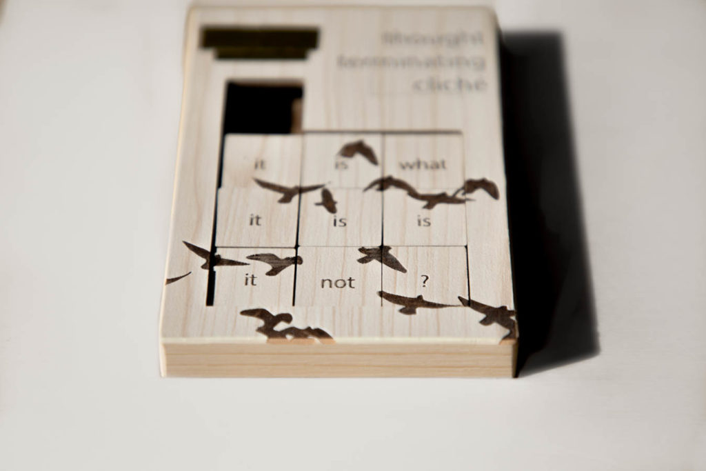 A poetic object called Thought-Terminating Cliché. A slide puzzle with a beige wooden texture lies on a white wooden table. The title of the piece is burned in the right corner but is too out of focus to read. The left corner has gold tape on it. The portion that slides is a 3x3 square. Each square contains a word, which reads "it is what it is is it not?" Also burned into squares are silhouettes of birds.