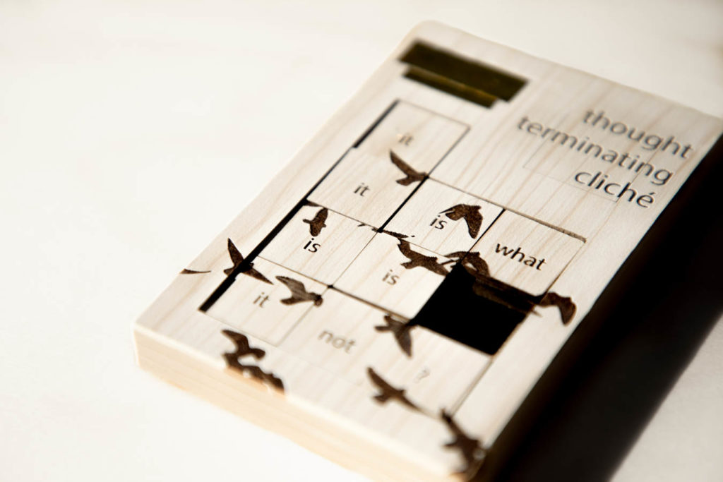 A poetic object called Thought-Terminating Cliché. A slide puzzle with a beige wooden texture lies diagonally on a white wooden table. The title of the piece is burned in the right corner. The left corner has gold tape on it. The portion that slides is a 3x3 square. Each square contains a word, which have been shuffled out of order. It reads "it it is what is is it not?" Also burned into squares are silhouettes of birds.