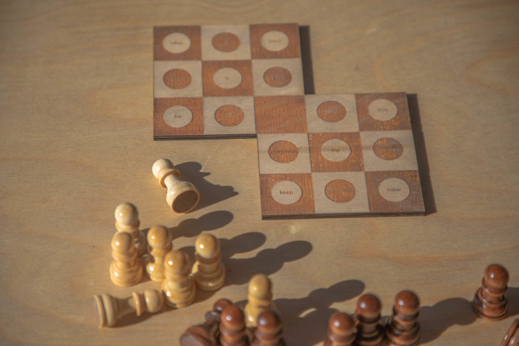 A photo of a poetry objected called White Flight. Two 3x3 checker-patterned boards are connected at one corner. On every square but that corner are circles with words burned in them. Most of the words are out of focus or too far away to read, but they include words like "keep," "becoming," and "mine." Below them, white and black pawns are arranged haphazardly, some spilled over. Their shadows are very long, cast on the wooden table.