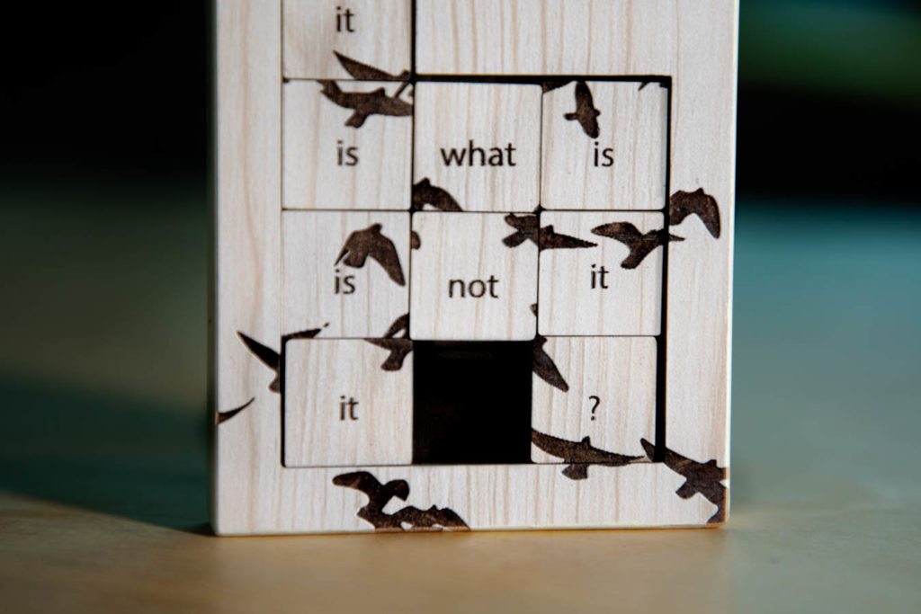 A close up of a poetic object called Thought-Terminating Cliché. A slide puzzle with a beige wooden texture stands on a wooden table. The visible portion of the puzzle is the portion that slides. It is a 3x3 square. Each square contains a word, which have been shuffled out of order. It reads "it is what is is not it it?" Also burned into squares are silhouettes of birds.
