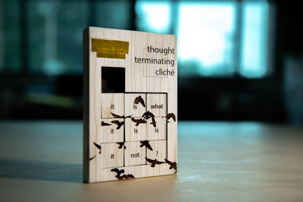 A poetic object called Thought-Terminating Cliché. A slide puzzle with a beige wooden texture stands on a wooden table. The title of the piece is burned in the right corner. The left corner has gold tape on it. The portion that slides is a 3x3 square. Each square contains a word, which reads "it is what it is is it not?" Also burned into squares are silhouettes of birds. In the background is an out of focus light source.