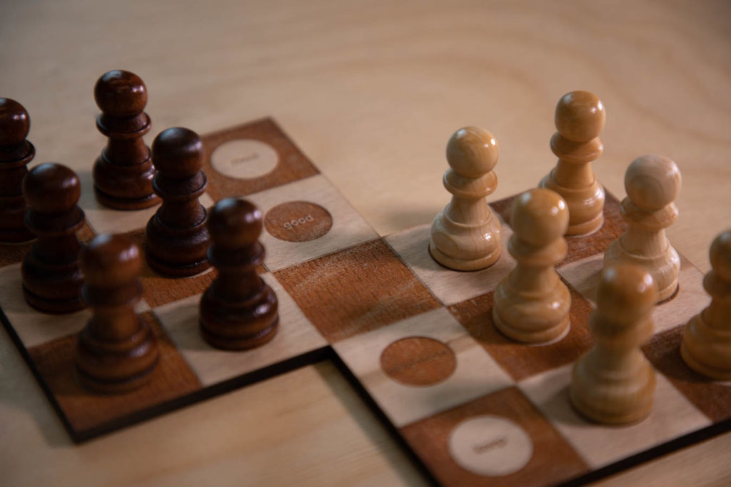 A close-up photo of a poetry objected called White Flight. Two 3x3 checker-patterned boards are connected at one corner. Most of the checkered squares have white pawns on them: black on one side and white on the other. A line of squares going down the center of the board have no pawns on them, and 4 of the 5 squares, have circles with words burned under them. The only one that is in focus is the word "good." Everything sits on a wooden table.