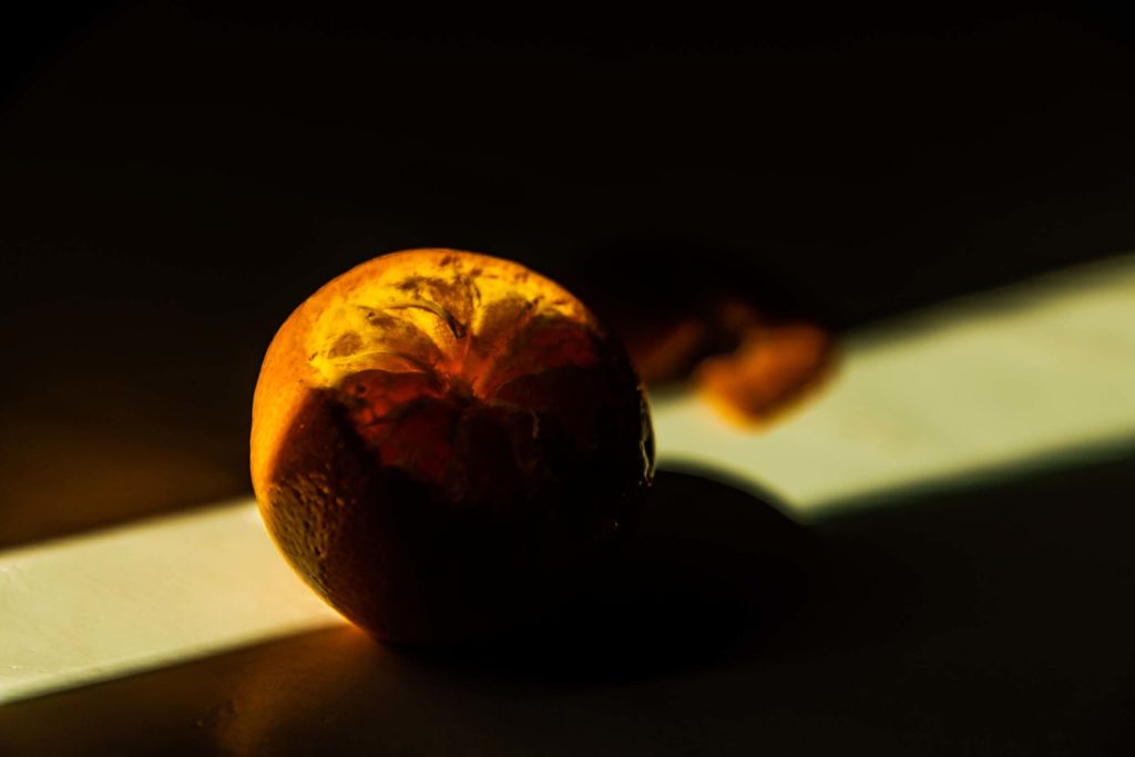 A photograph of an orange, partially peeled. A diagonal line of light cuts behind, and partially over the orange, causing the flesh to seem to glow. In the light, out of focus, are the remaining peels. The shadows above and below the orange make it too dark to make anything else out.